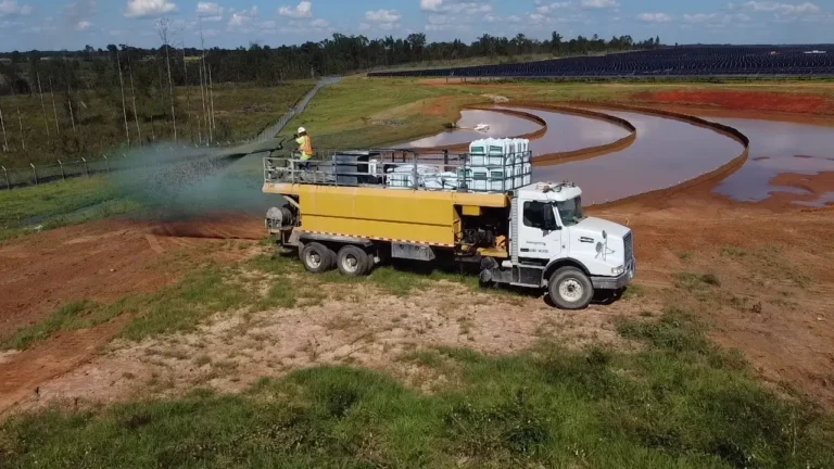 A yellow truck spraying green liquid for erosion control on a grassy slope near a water retention pond at a solar farm, with a worker standing on the truck bed.
