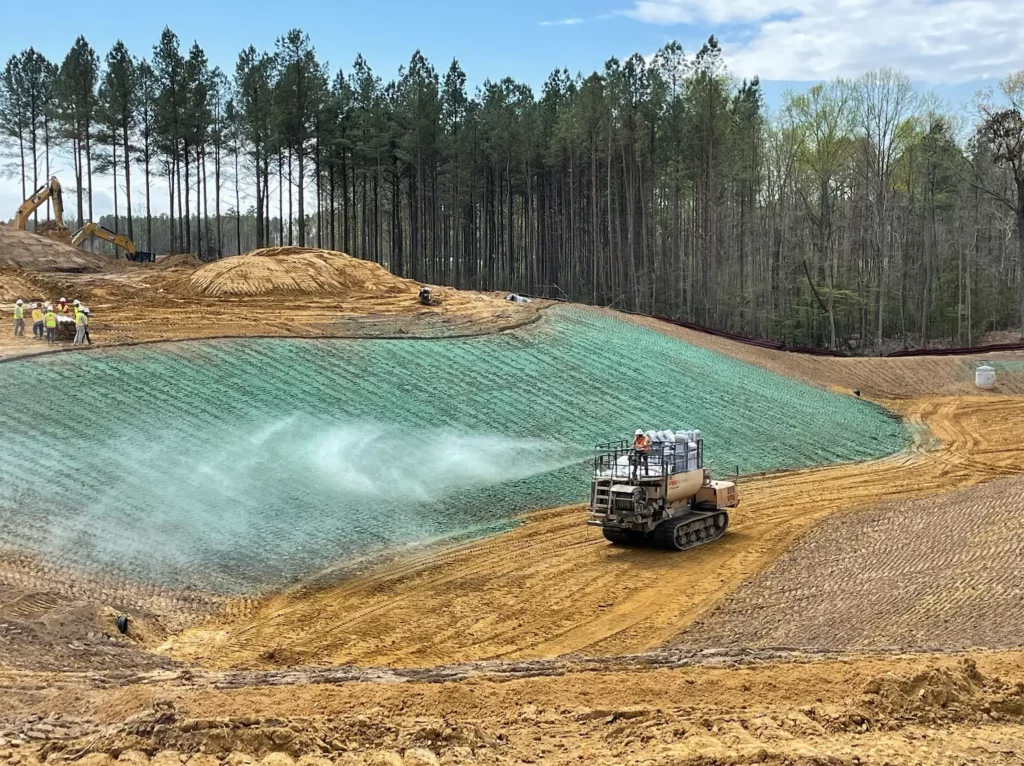 Tracked hydroseeding vehicle spraying green slurry on a dirt slope, surrounded by construction workers, excavators, and dense forest under a partly cloudy sky.