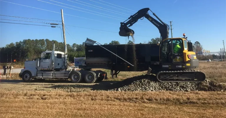 An excavator loads crushed rocks into a dump truck trailer at a roadside construction site with power lines, grass, and trees in the background.