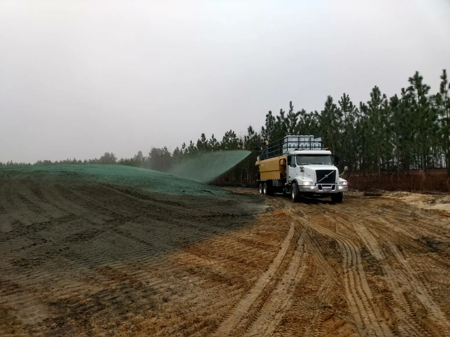 A truck sprays green hydroseeding mixture on a large, sloped area covered with erosion control mesh beside a dirt road under a clear blue sky.