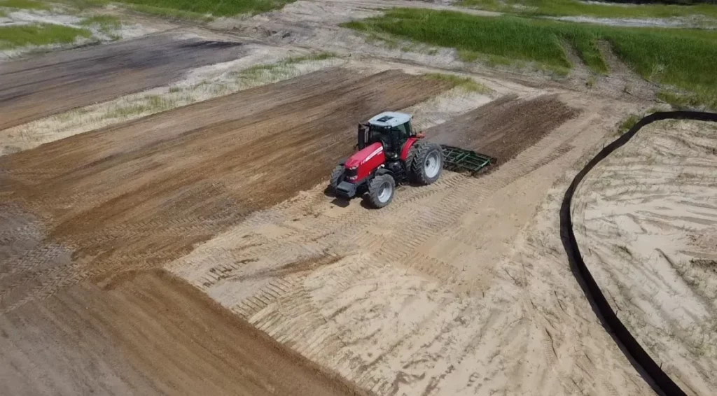 A red tractor with a soil cultivator prepares a graded sandy construction site, surrounded by sparse vegetation and erosion control barriers.