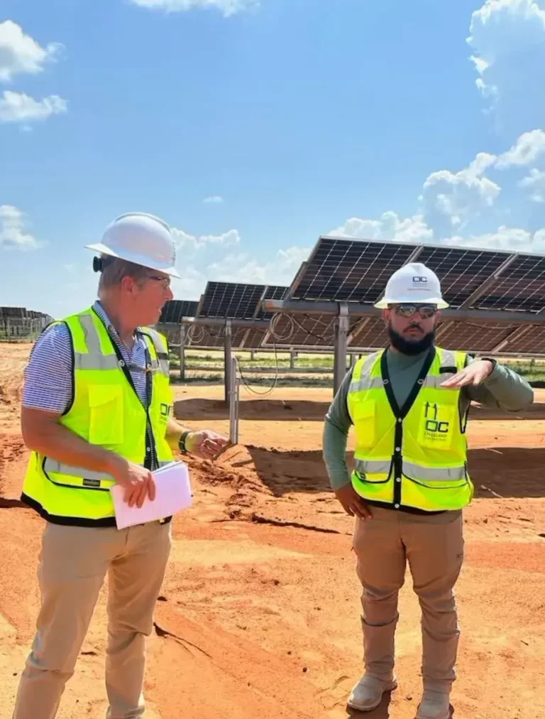 Two men in high-visibility vests and hard hats discussing near solar panels on a sandy construction site under a sunny sky.