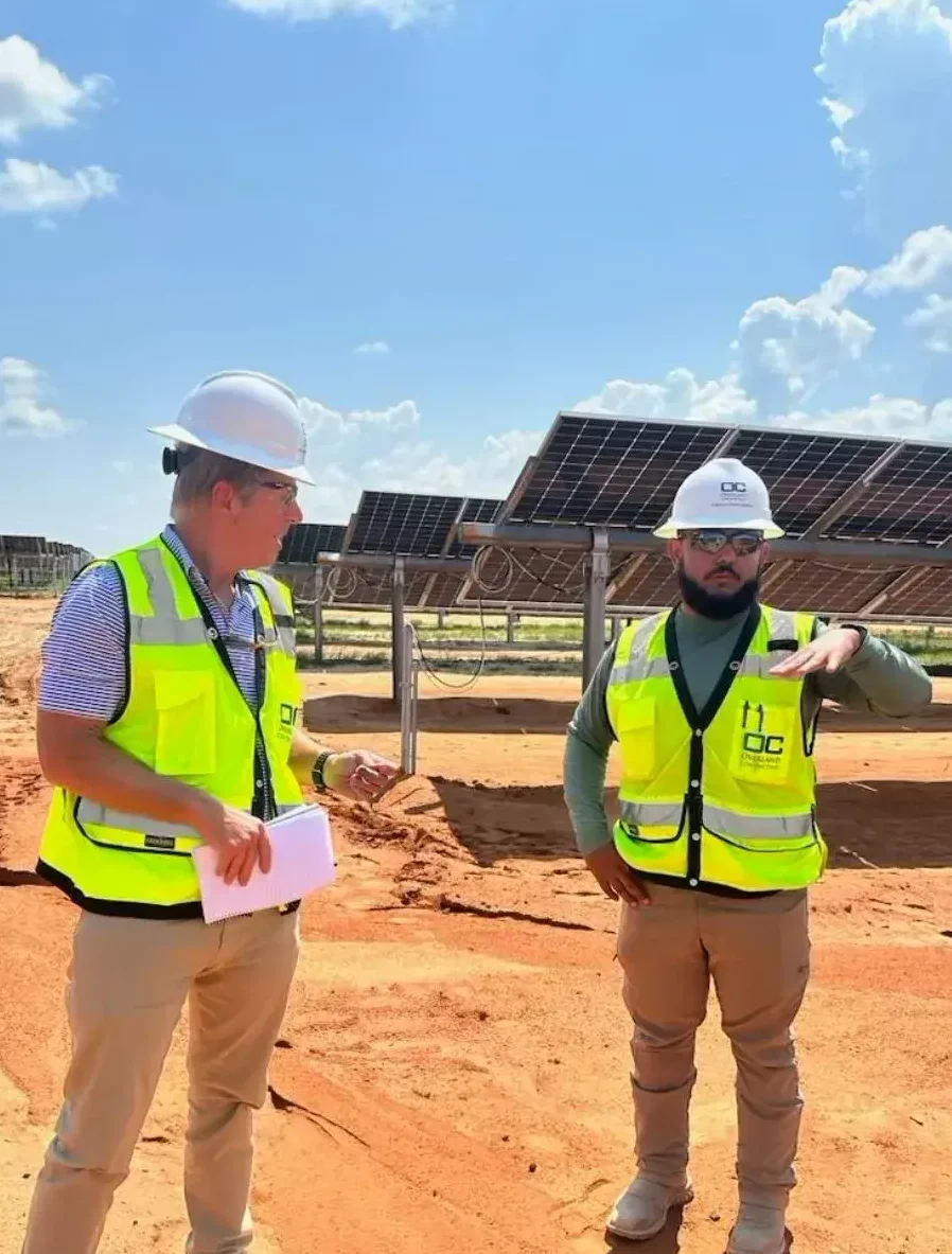 Two men in high-visibility vests and hard hats discussing near solar panels on a sandy construction site under a sunny sky.